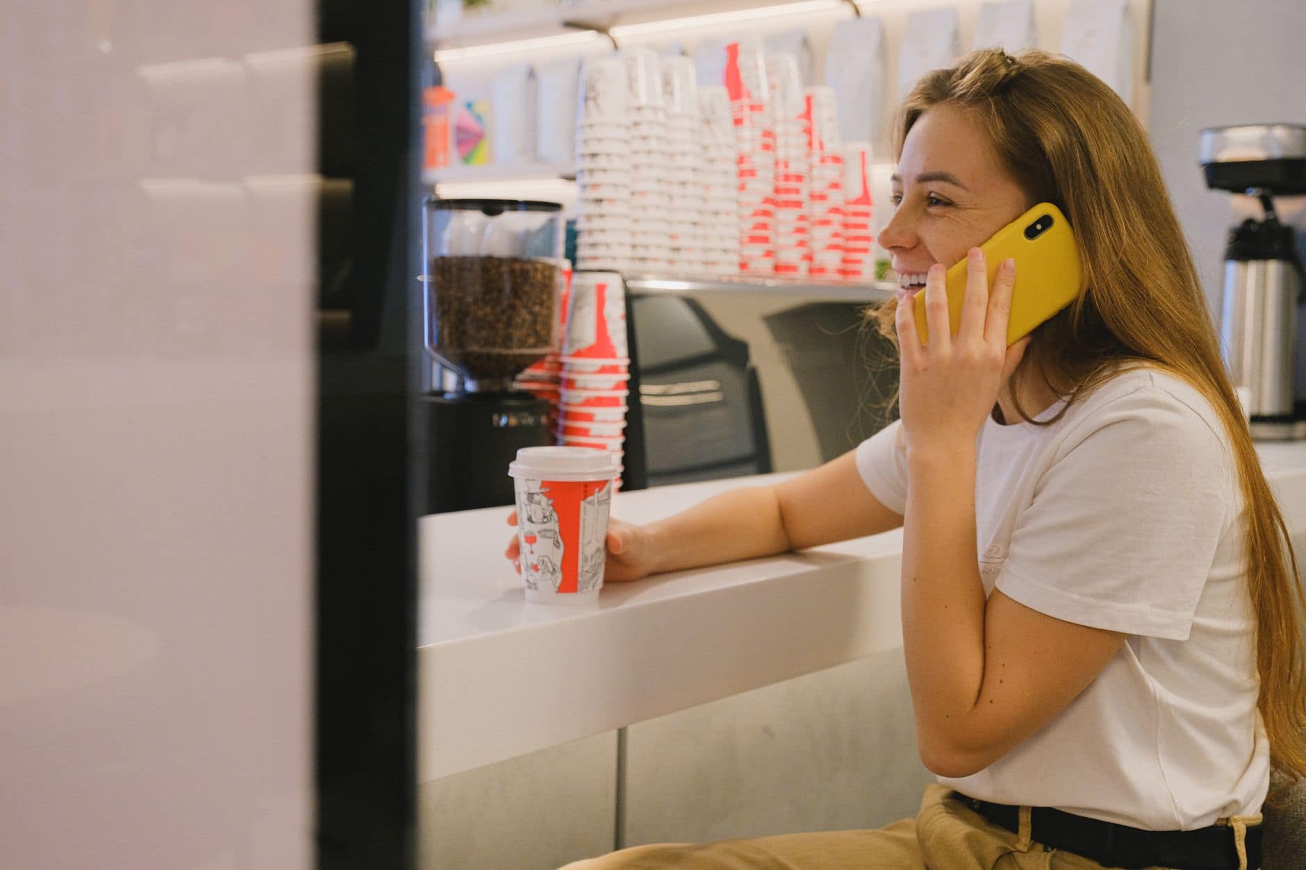 Una donna sorride mentre parla al telefono e tiene un bicchiere di caffè al bancone.