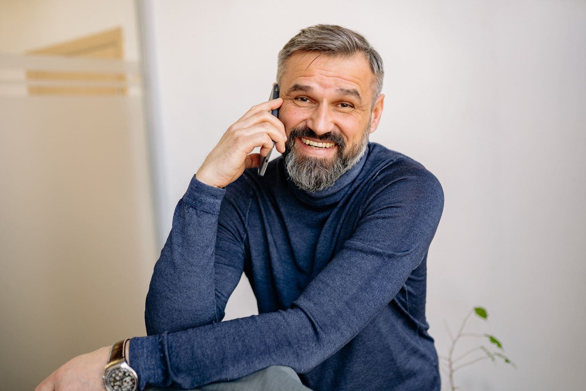 Um homem de barba e camisola azul sorri com um telemóvel ao ouvido.