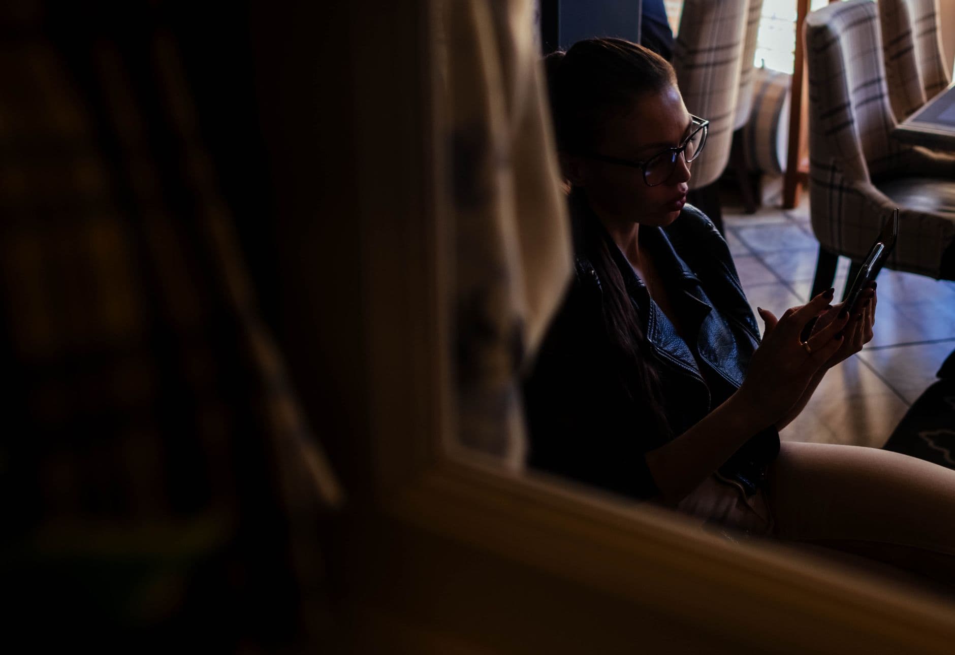 Een jonge vrouw met een bril zit in een schemerige ruimte en leest iets op haar smartphone.