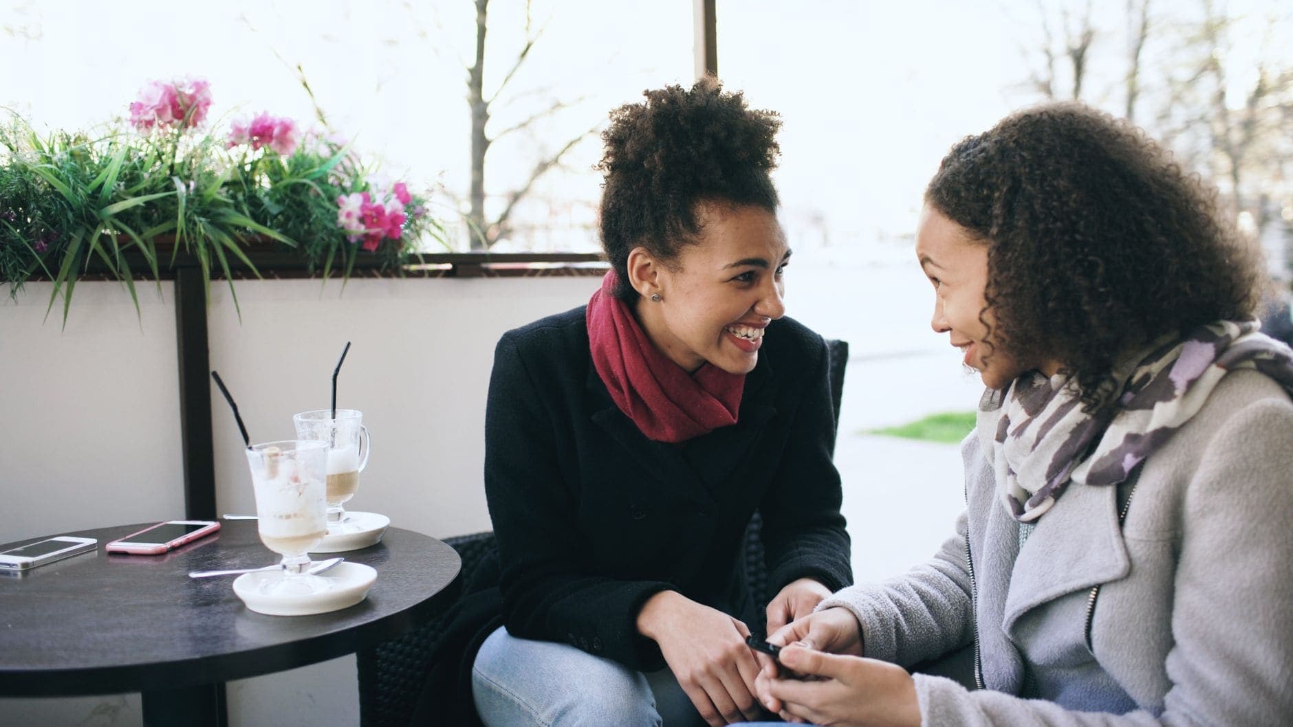 Two women in coats and scarves chat beside a café table with tall coffee glasses.