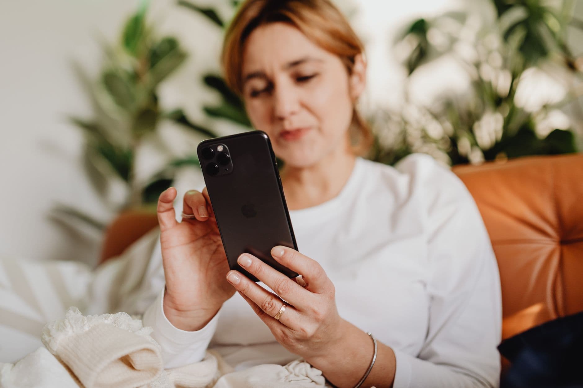 Een vrouw in een witte blouse gebruikt haar telefoon op een bruine zetel, naast kamerplanten.