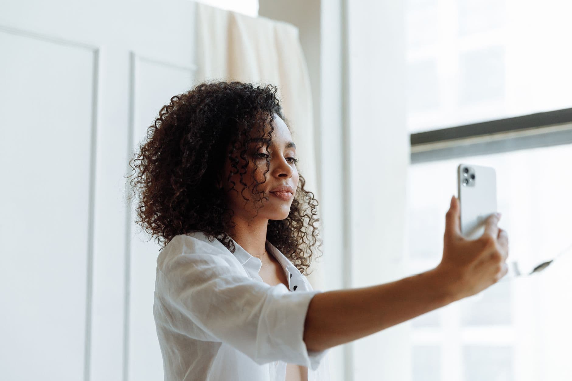 Eine Frau mit lockigem Haar macht am hellen Fenster ein Selfie mit ihrem Smartphone.
