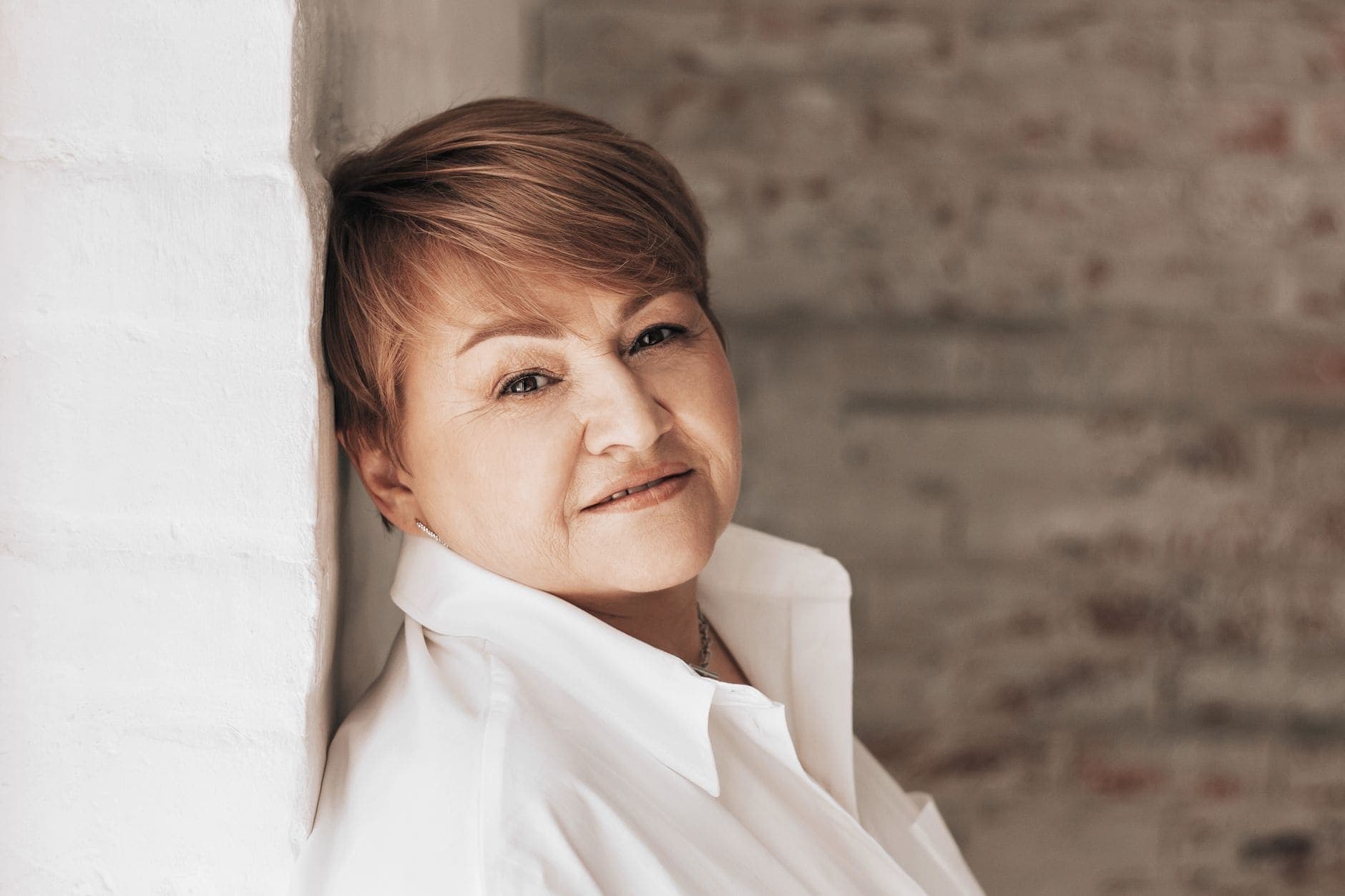 A woman in a white blouse leans against a pale wall and looks towards the camera.