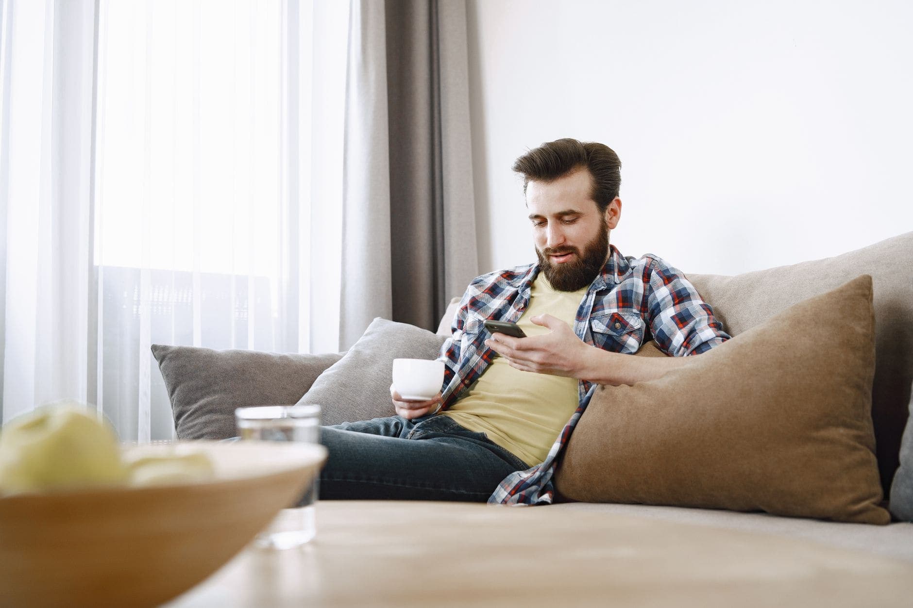 A man in a checked shirt sits on a sofa, holding a white cup and looking at his phone.