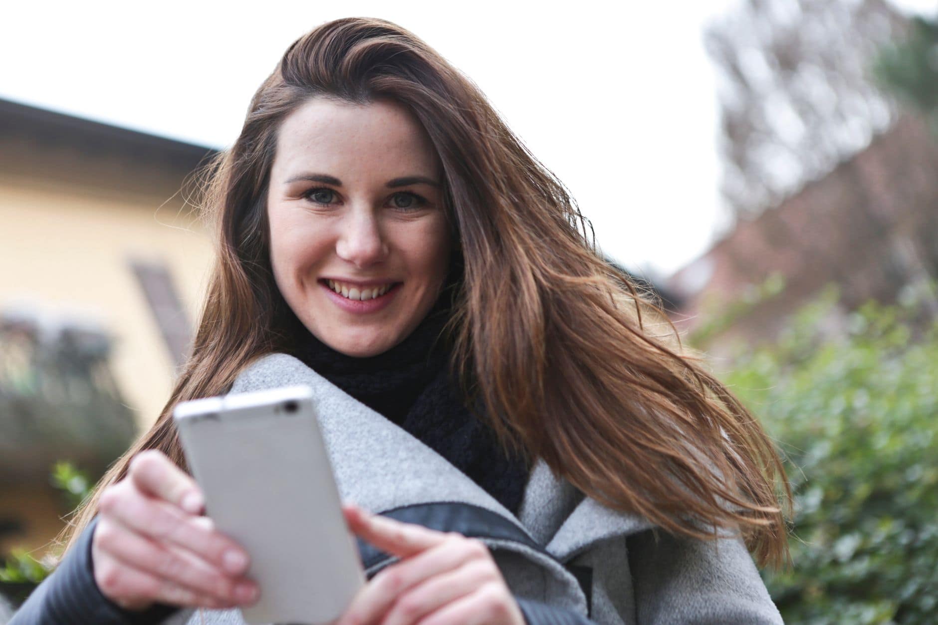 Eine lächelnde Frau schaut im Freien auf ihr Smartphone, das sie mit beiden Händen hält.