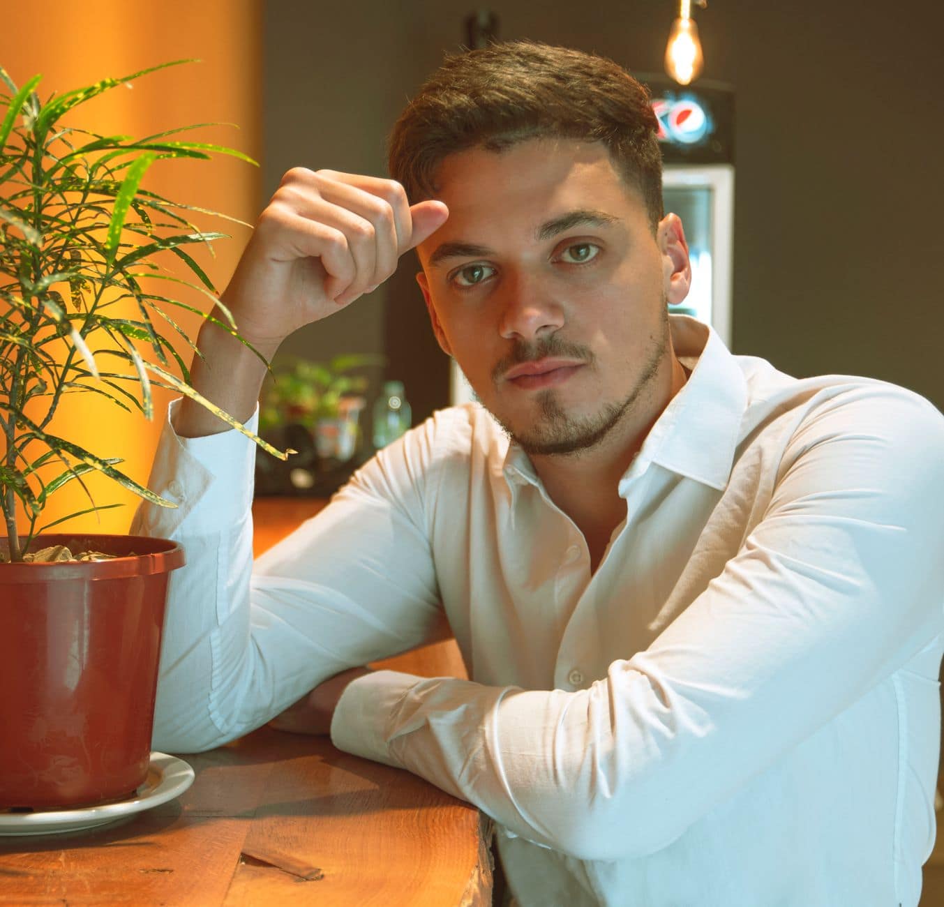 Muž s modrými očami v bielej košeli sedí pri drevenom stole s rukou pri čele