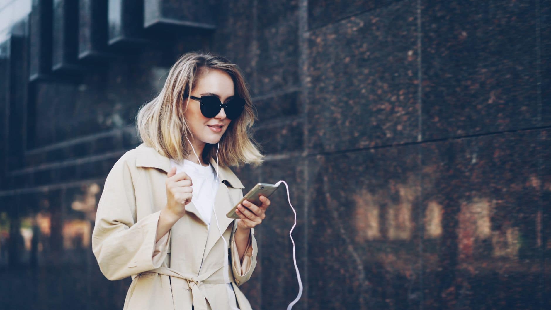 Kobieta w beżowym płaszczu trzyma telefon ze słuchawkami na tle ceglanej ściany