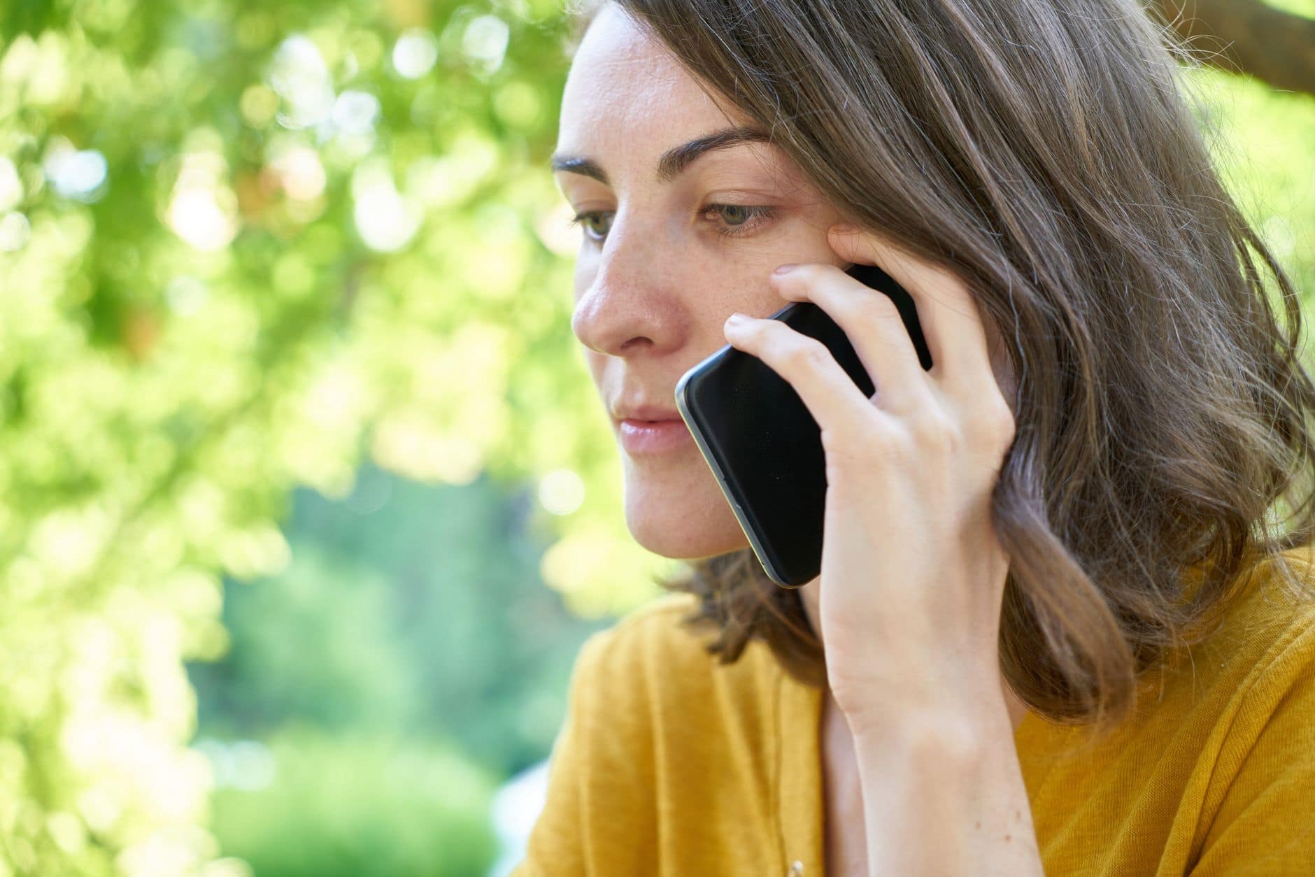 Kobieta w żółtej koszulce rozmawia przez telefon wśród zielonych drzew