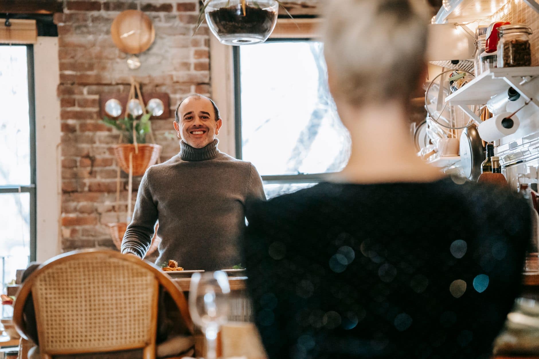 Un homme en pull gris sourit à une femme vue de dos, près d'une table.