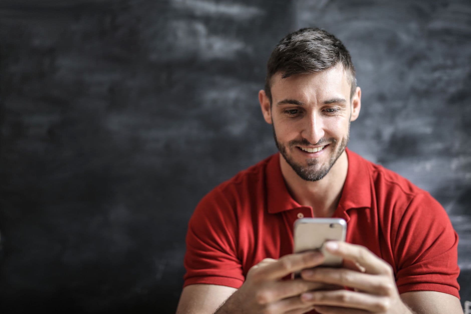 Un uomo sorridente in polo rossa guarda lo schermo del suo smartphone su sfondo scuro.