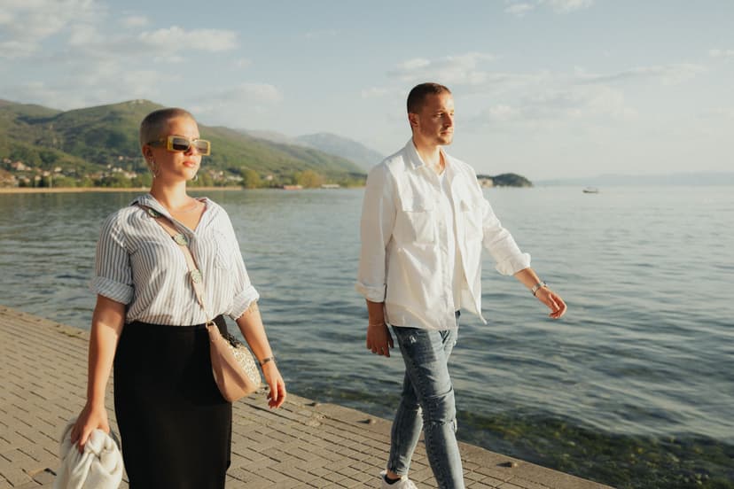 Et stilfullt par går langs en solfylt strandpromenade ved sjøen i varmt kveldslys, hun i stripete skjorte og solbriller, han i hvit skjorte, med fjell i bakgrunnen