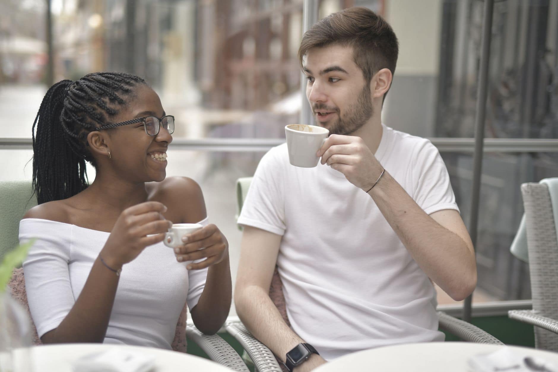 Een vrouw met een bril en een man in een wit T-shirt praten glimlachend met koffie in de hand.