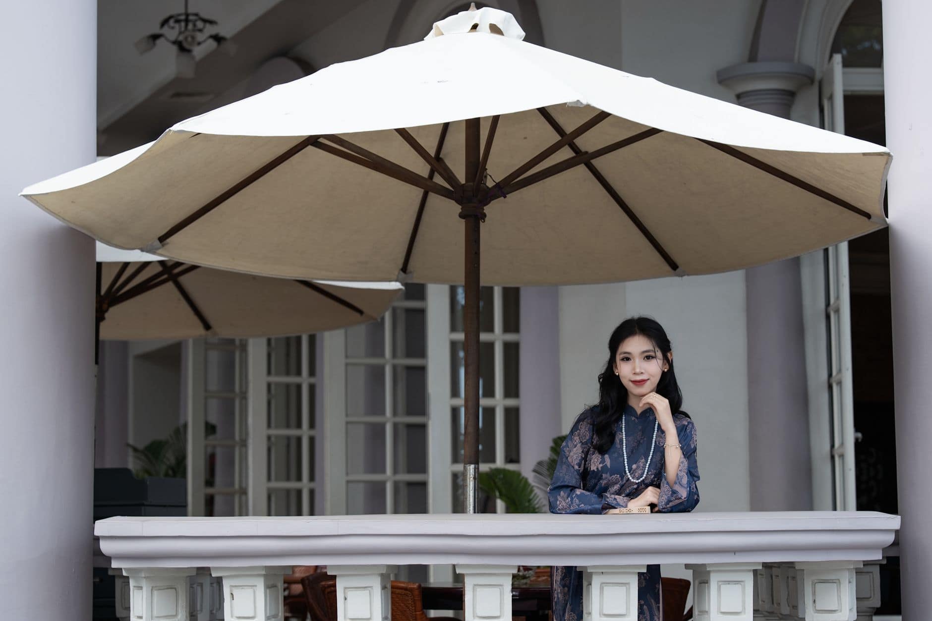 Une femme en robe bleue se tient sous un grand parasol blanc sur une terrasse.
