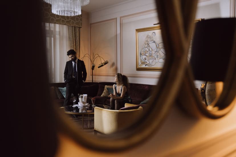 Elegant par i en klassisk hotellsvit, där mannen i mörk kostym står vid soffgruppen medan kvinnan i paljettklänning sitter i soffan med champagne och glas framdukade på bordet