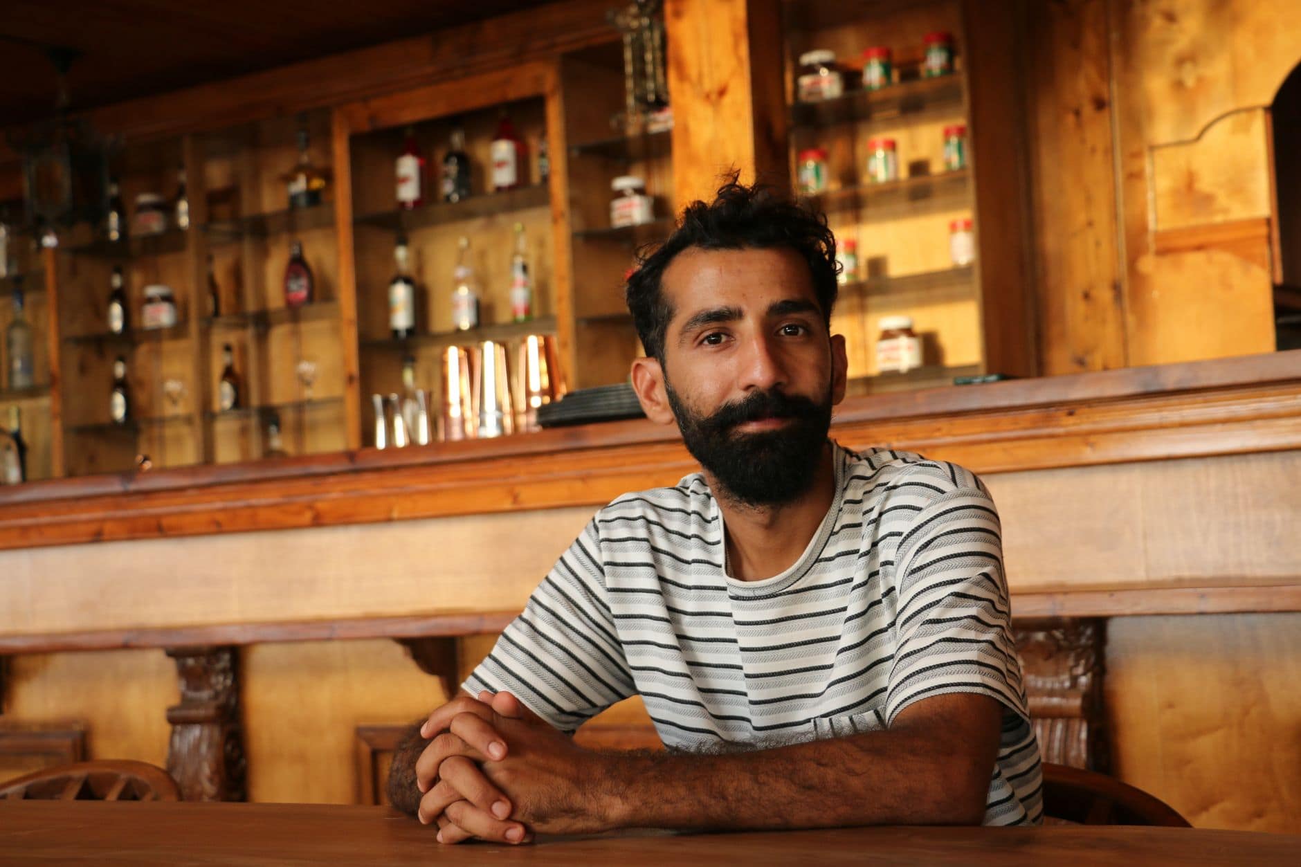 Un uomo con la barba e una maglietta a righe siede con le mani intrecciate davanti a un bancone di legno.