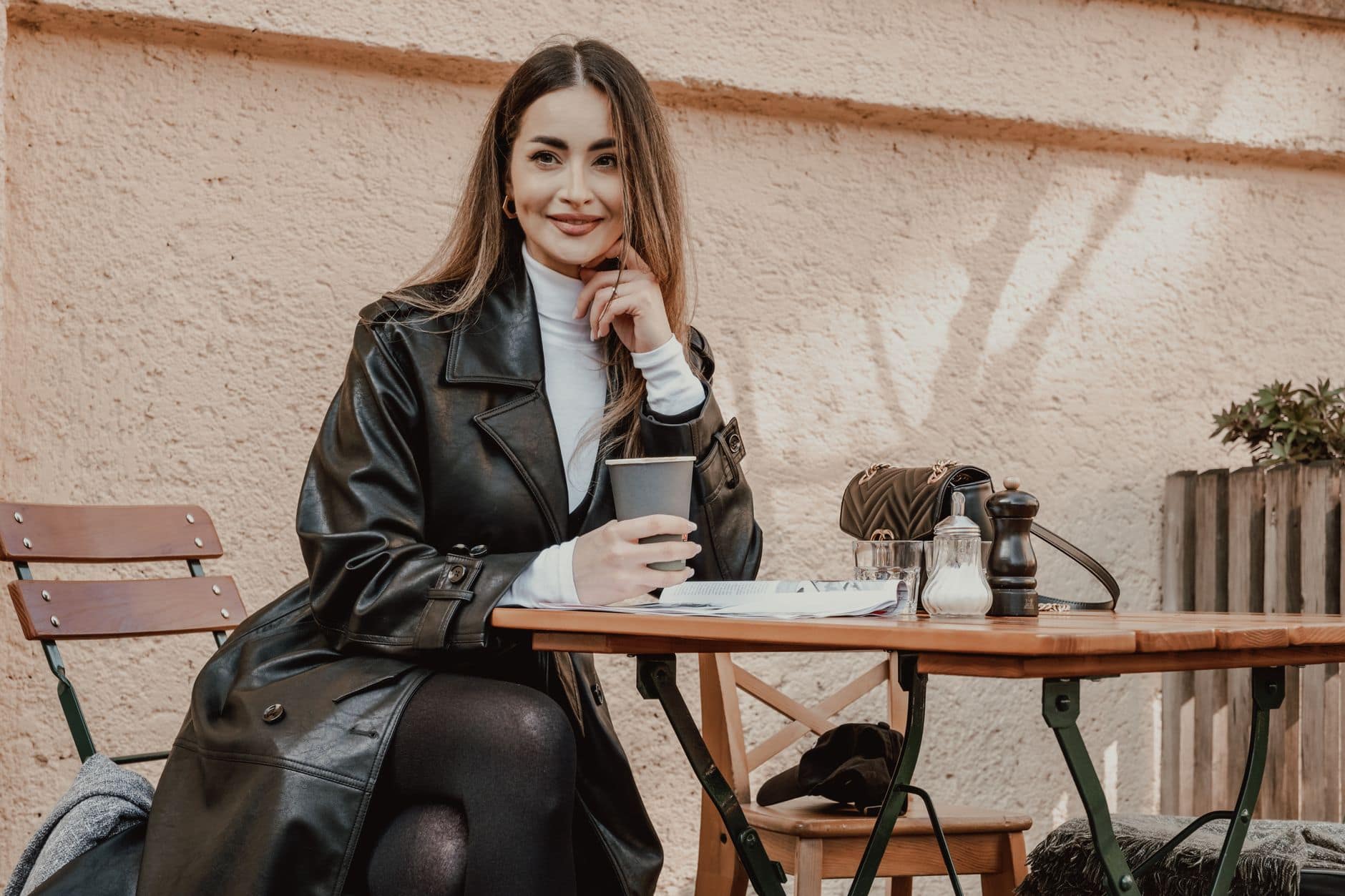 Una donna con un cappotto nero sorride al tavolo di un bar tenendo un bicchiere di caffè.