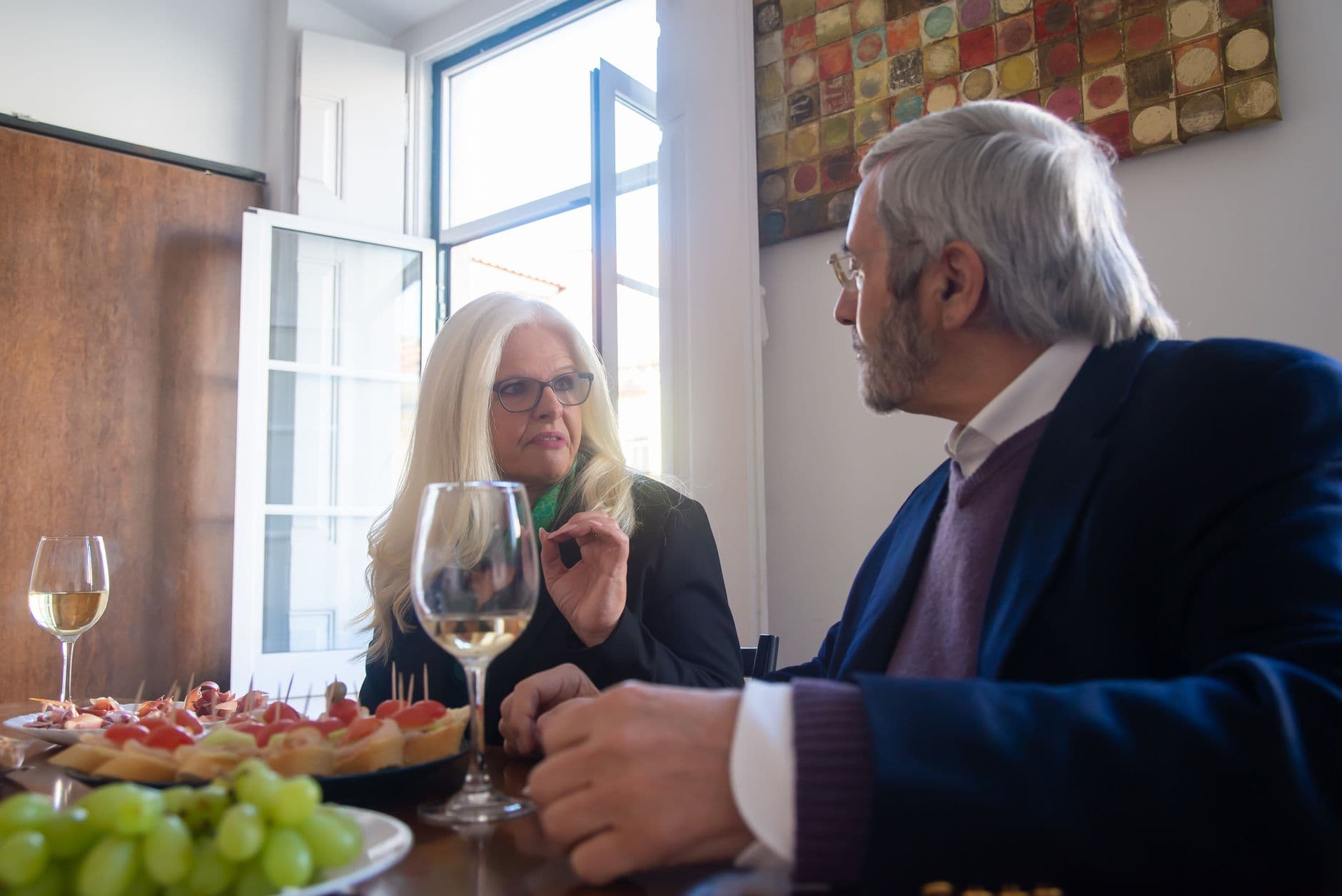 Un homme et une femme aux cheveux blancs discutent à table devant des verres de vin blanc.