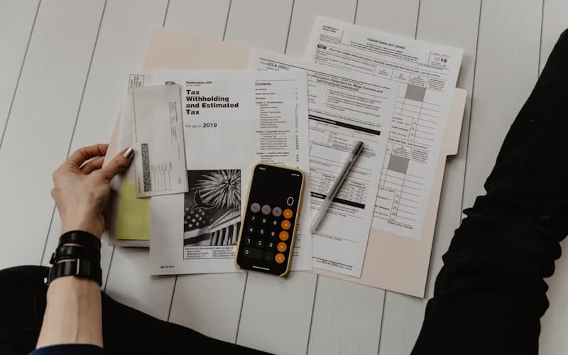 Overhead view of a hand among tax forms, a pen and a phone calculator on white floorboards.
