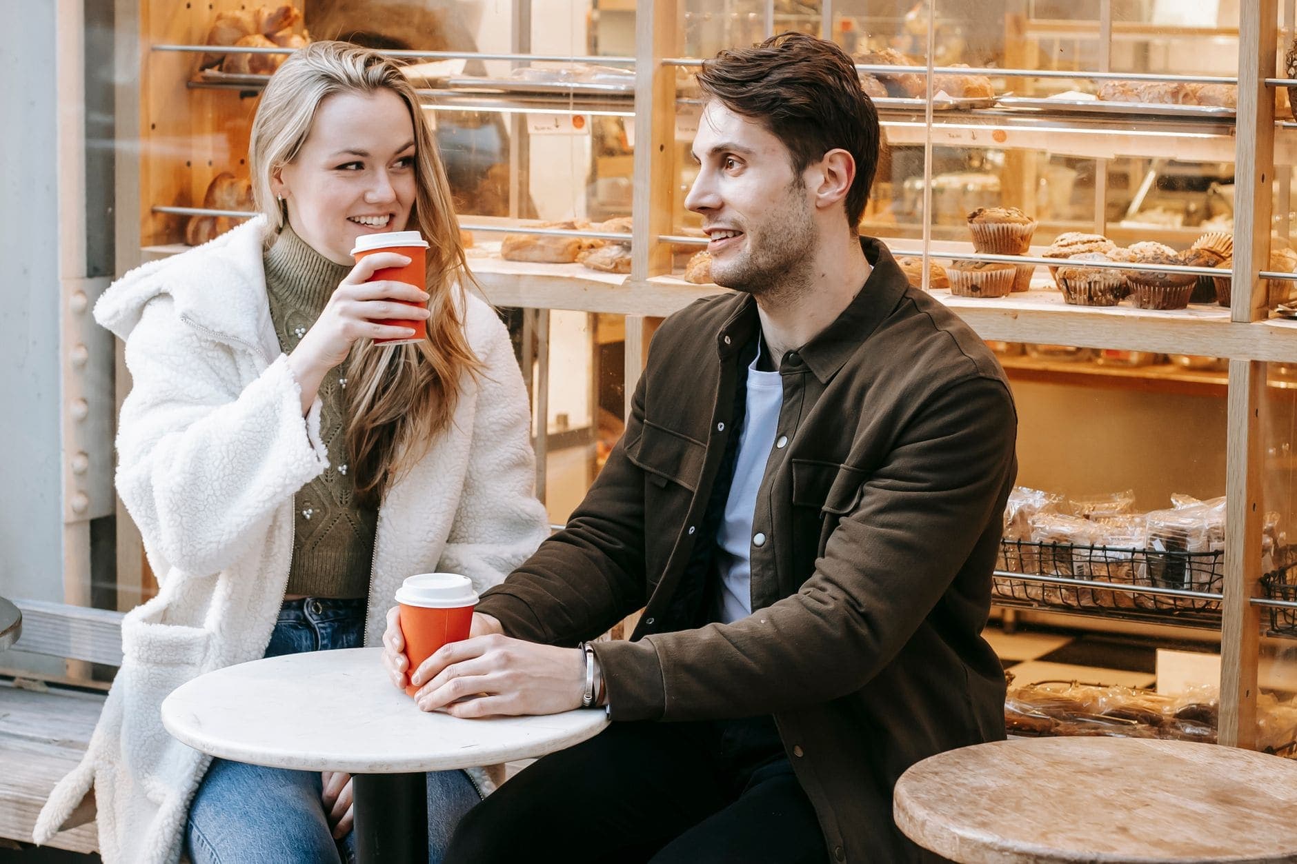 Uma mulher e um homem sorriem um para o outro numa mesa junto à montra de uma pastelaria.