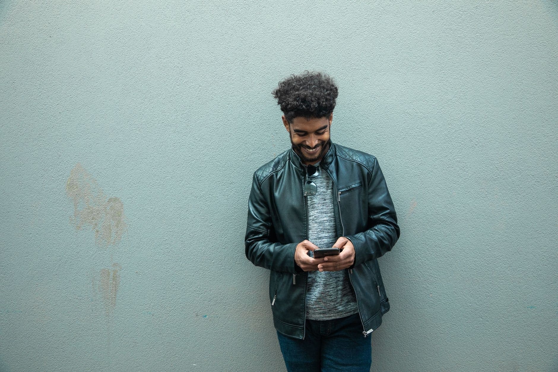 Um homem de jaqueta escura sorri ao olhar para o celular, parado diante de uma parede clara.