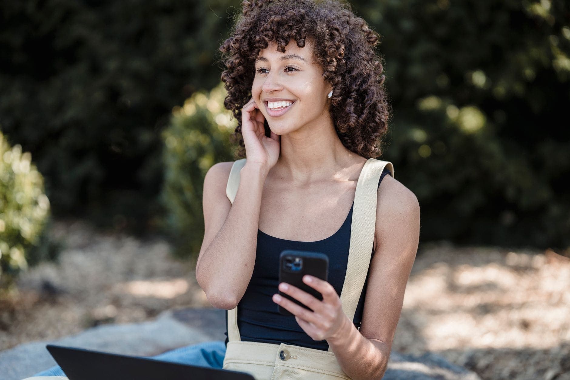 Uma mulher de cabelo cacheado sorri para o lado, segurando um celular ao ar livre.