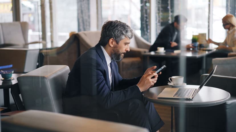 Elegant gråhårig man i mörkblå kostym sitter avslappnat i en stilfull kafélounge och läser ett meddelande på sin smartphone, med en kaffekopp på bordet