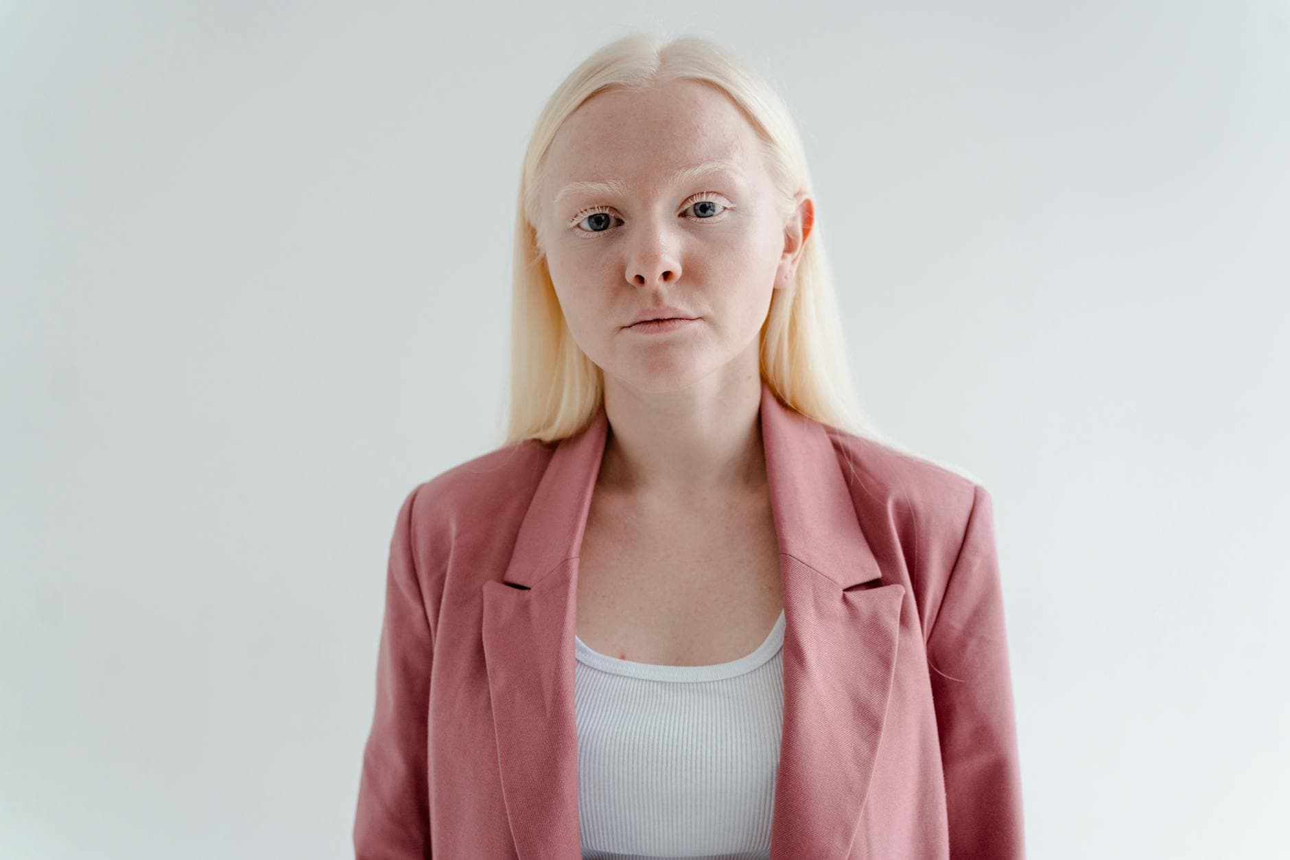 Person med mycket ljust hår i rosa kavaj och vitt linne mot en ljusgrå bakgrund.