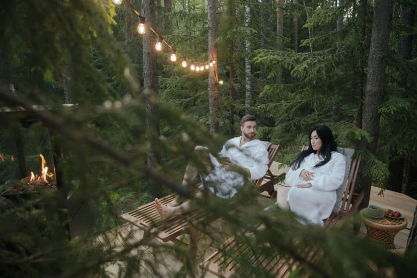 Pari valkoisissa kylpytakeissa rentoutuu vierekkäin aurinkotuoleissa mökin puuterassilla keskellä havumetsää - valosarja hehkuu puiden välissä, tulisija loimuaa vieressä ja kahvit on katettu pikkupöydälle