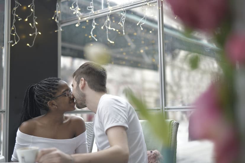 A young couple leans in for a kiss over coffee at a café window table strung with fairy lights, with the grey winter street visible through the glass behind them