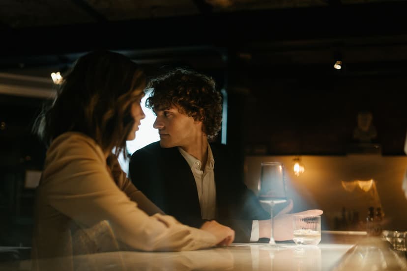 A well-dressed couple talks closely over a glass of red wine at a softly lit bar counter, the man in a dark suit listening intently to his companion