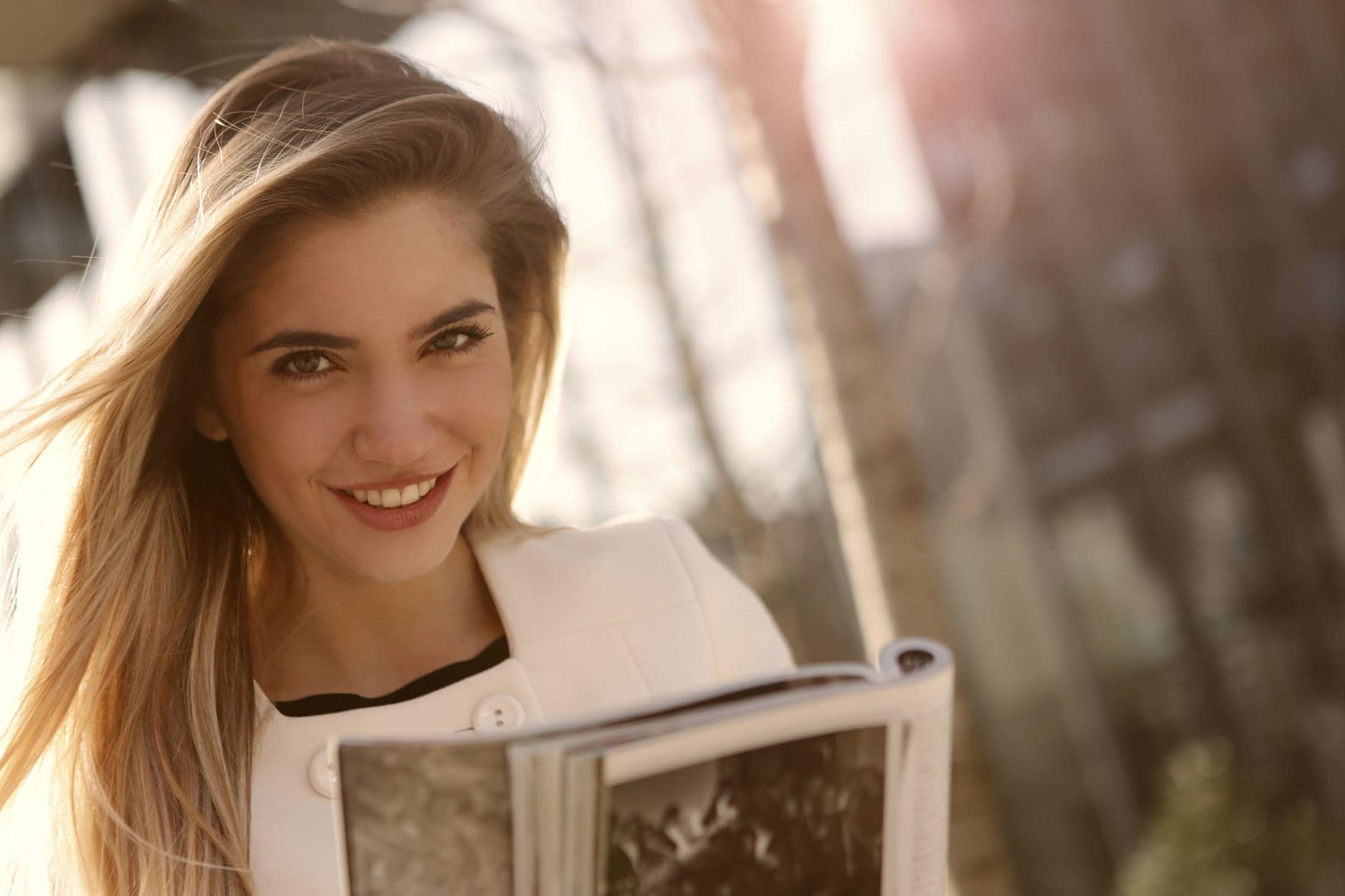 Une femme souriante en veste blanche tient un magazine ouvert en plein air.