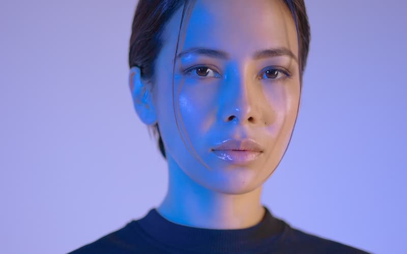 Studio portrait of a woman with dark hair in a black top under blue light on a lilac background.