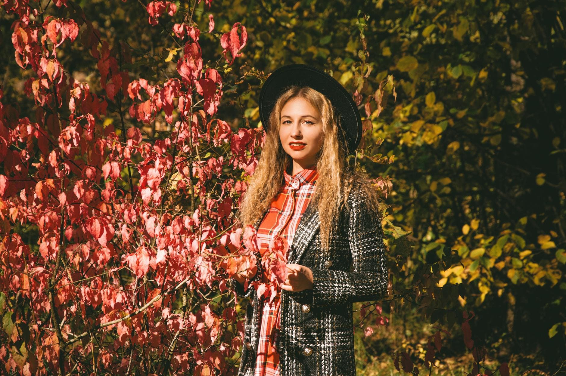 A woman in a black hat and grey tweed coat stands beside a bush of red autumn leaves.