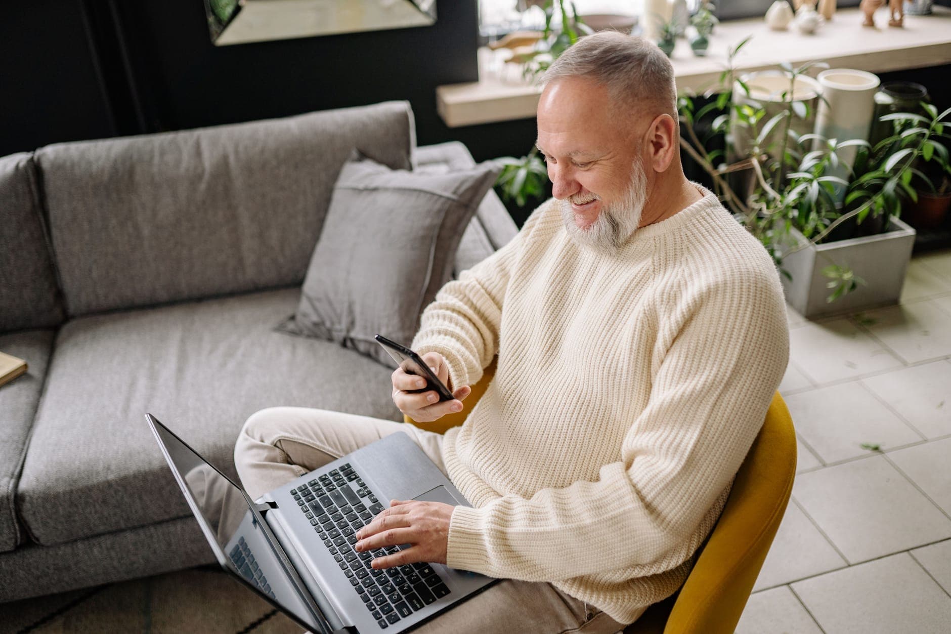 A smiling gray-bearded man in a cream sweater uses a laptop and a phone on a chair in his living room.