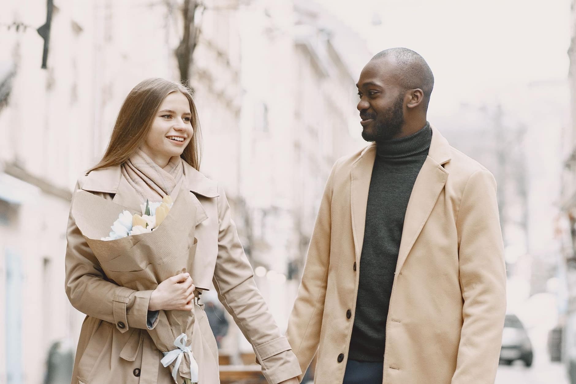 Smiling woman with a bouquet walks hand in hand with a man in a camel coat down a pale city street.