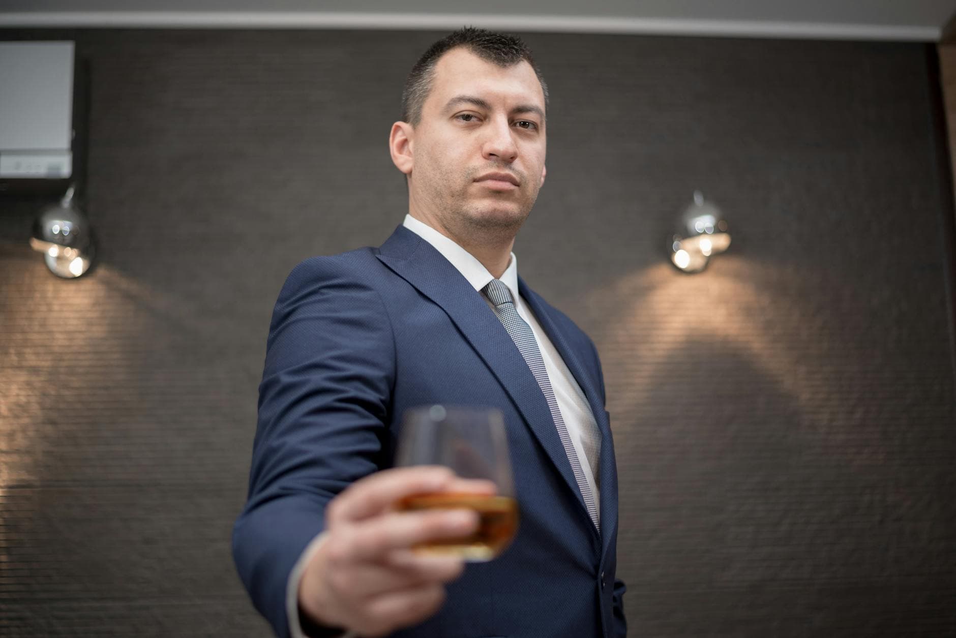 Mann im dunkelblauen Anzug hält ein Glas Whisky vor eine graue Wand mit Wandleuchten.