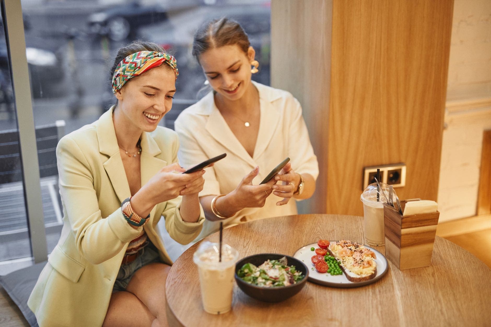 Duas mulheres sorriem, cada uma com seu celular, à mesa de um café.