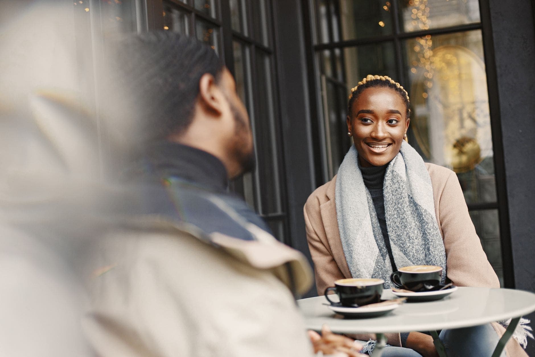A woman in a coat smiles at the person across a small café table with two coffees.