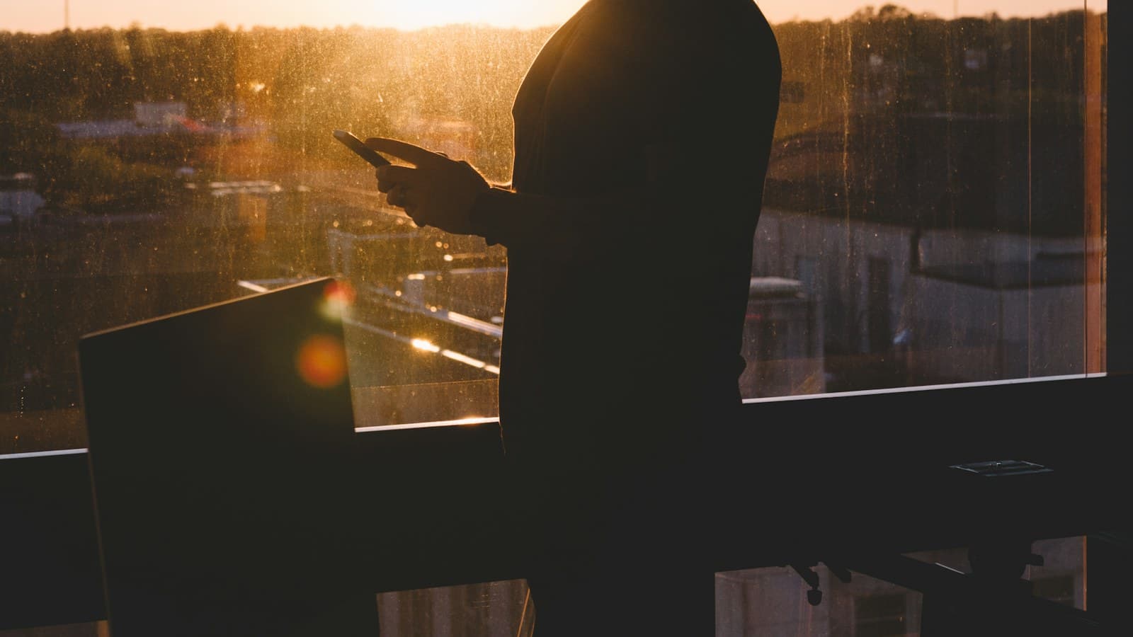Silhouette of a person holding a phone by a sunlit window at sunset, face out of frame