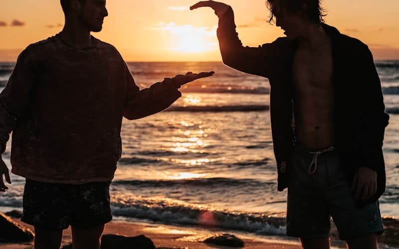 Två personer i motljus på en strand jämför längd med händerna medan solen går ner.