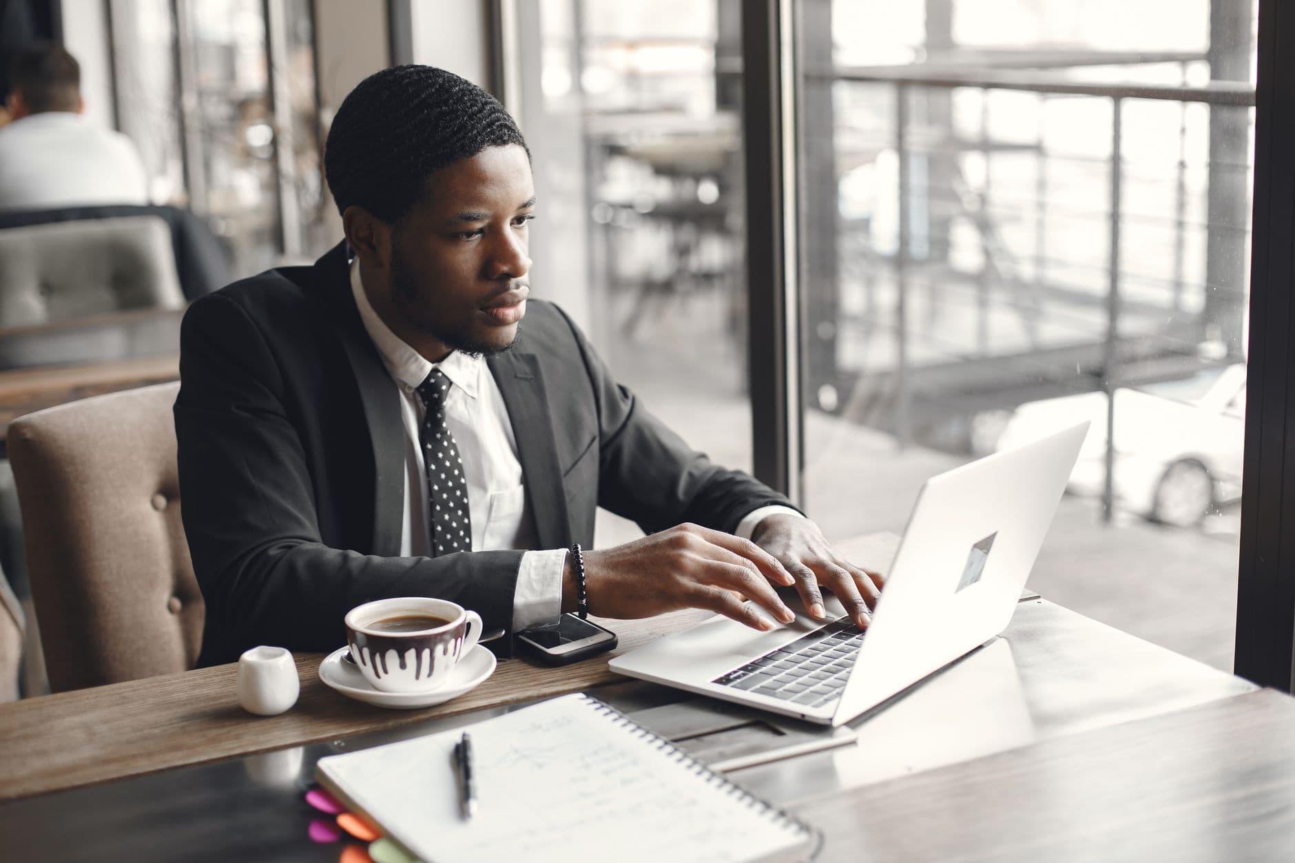 A man in a dark suit types on a laptop at a café table by the window, with coffee and a notebook beside him.