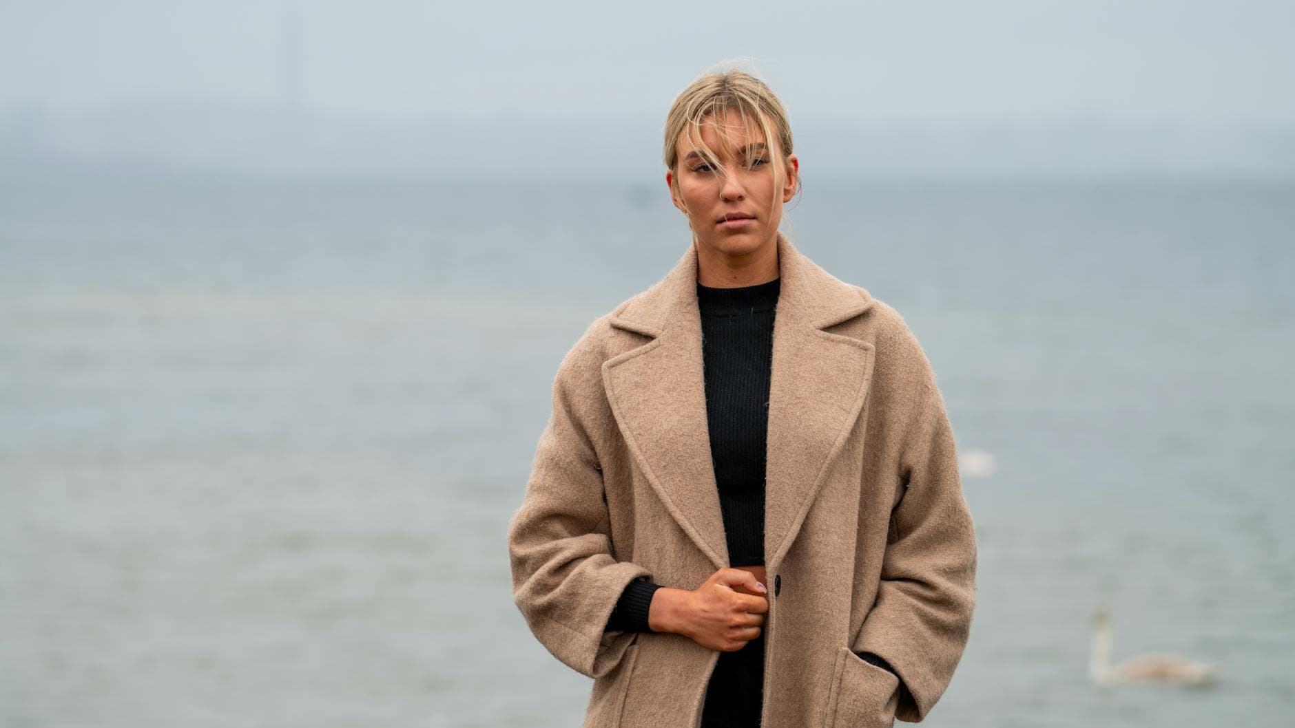 Lyshåret kvinde i beige uldfrakke står ved gråt vand med en svane i baggrunden.