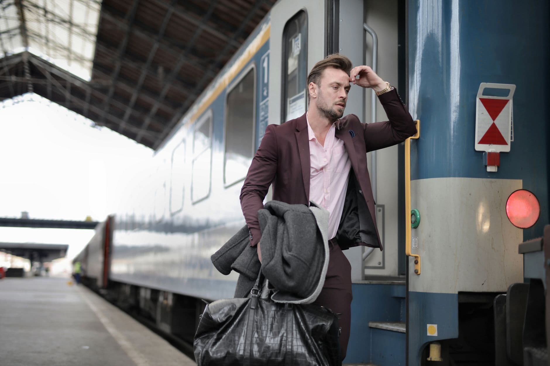 Een man in een bordeauxrode jas staat met een tas en kleding op een perron naast een trein.