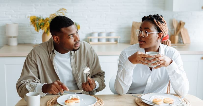 A man in an olive shirt talks with a woman holding a coffee mug over breakfast plates in a bright white kitchen.