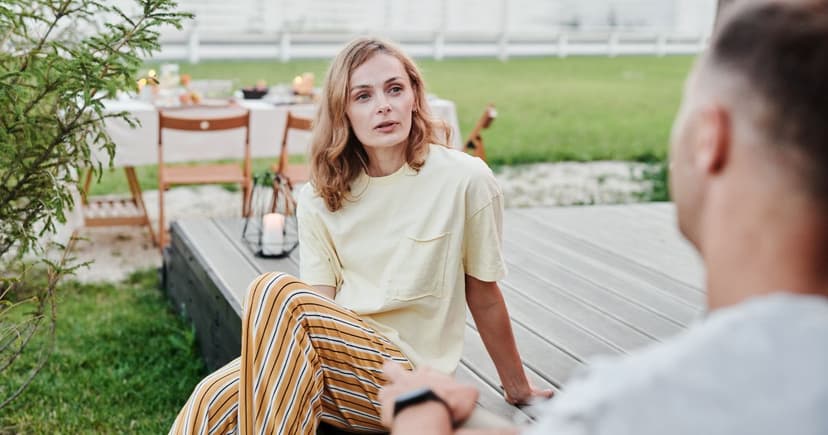 A woman in a pale yellow shirt sits on a wooden deck talking with a man, a candlelit garden table behind them.
