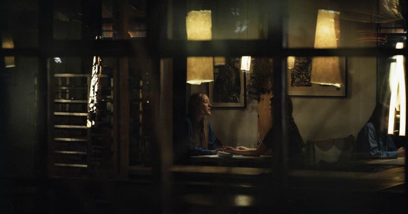 A woman in a blue shirt and her companion sit across a small table in a dim restaurant, hands almost meeting under low paper lamps