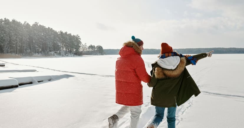 A woman in a red down parka and a man in a fur-hooded parka hold hands as they cross a snow-covered lake in knit hats and heavy boots, one of them pointing toward the far shore