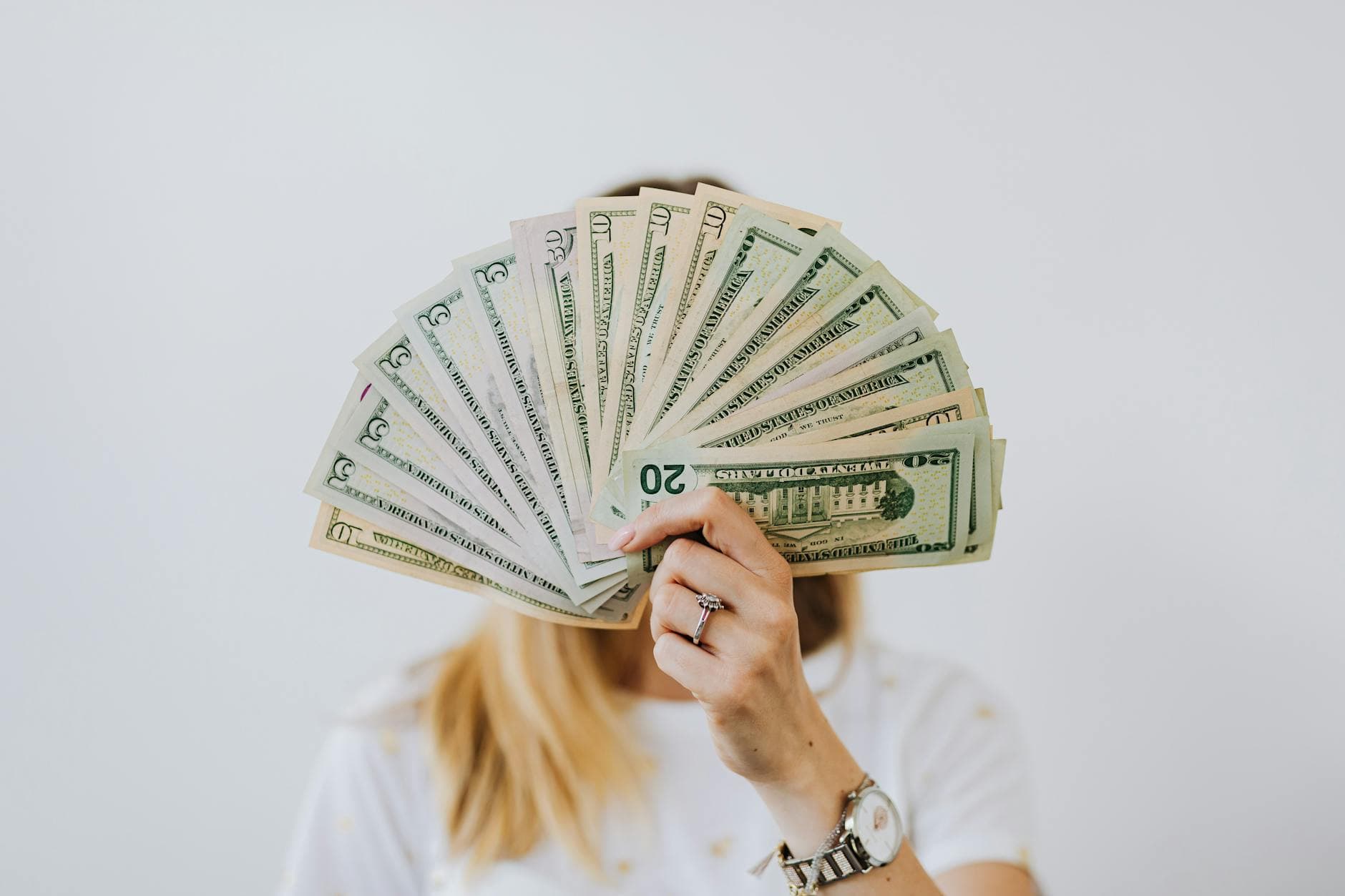 A hand holds a fan of banknotes in front of a face, against a pale grey wall.