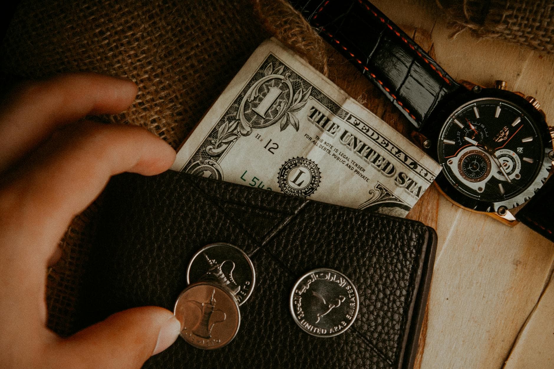 A hand reaches for a black leather wallet with a dollar bill, coins and a watch.