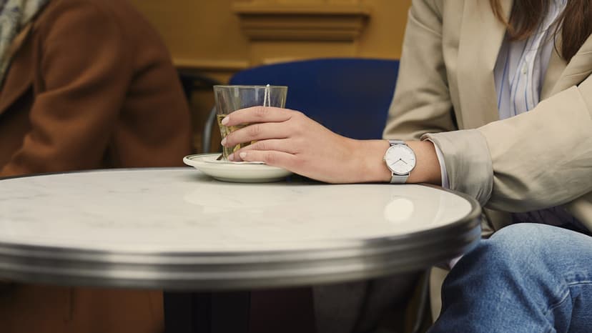 Two people in autumn coats at a small marble café table, one resting a hand on a glass of tea on its saucer, faces out of frame